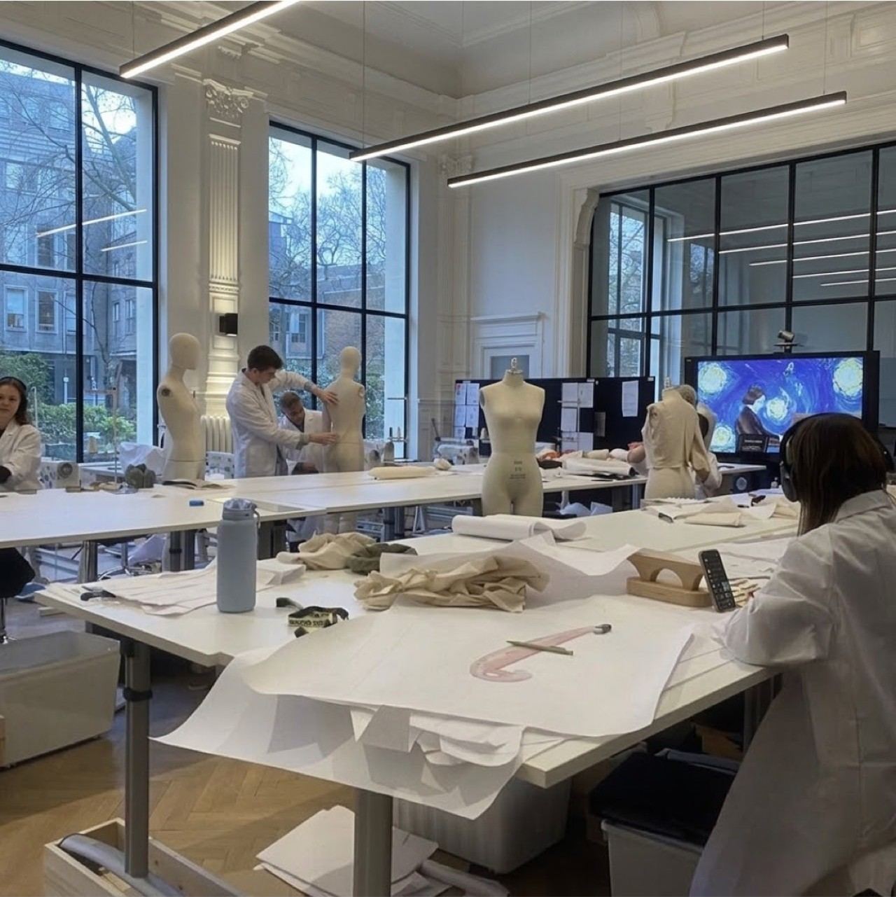 A light-filled fashion design studio: students draping and pattern cutting on dress forms at long white worktables beneath tall windows