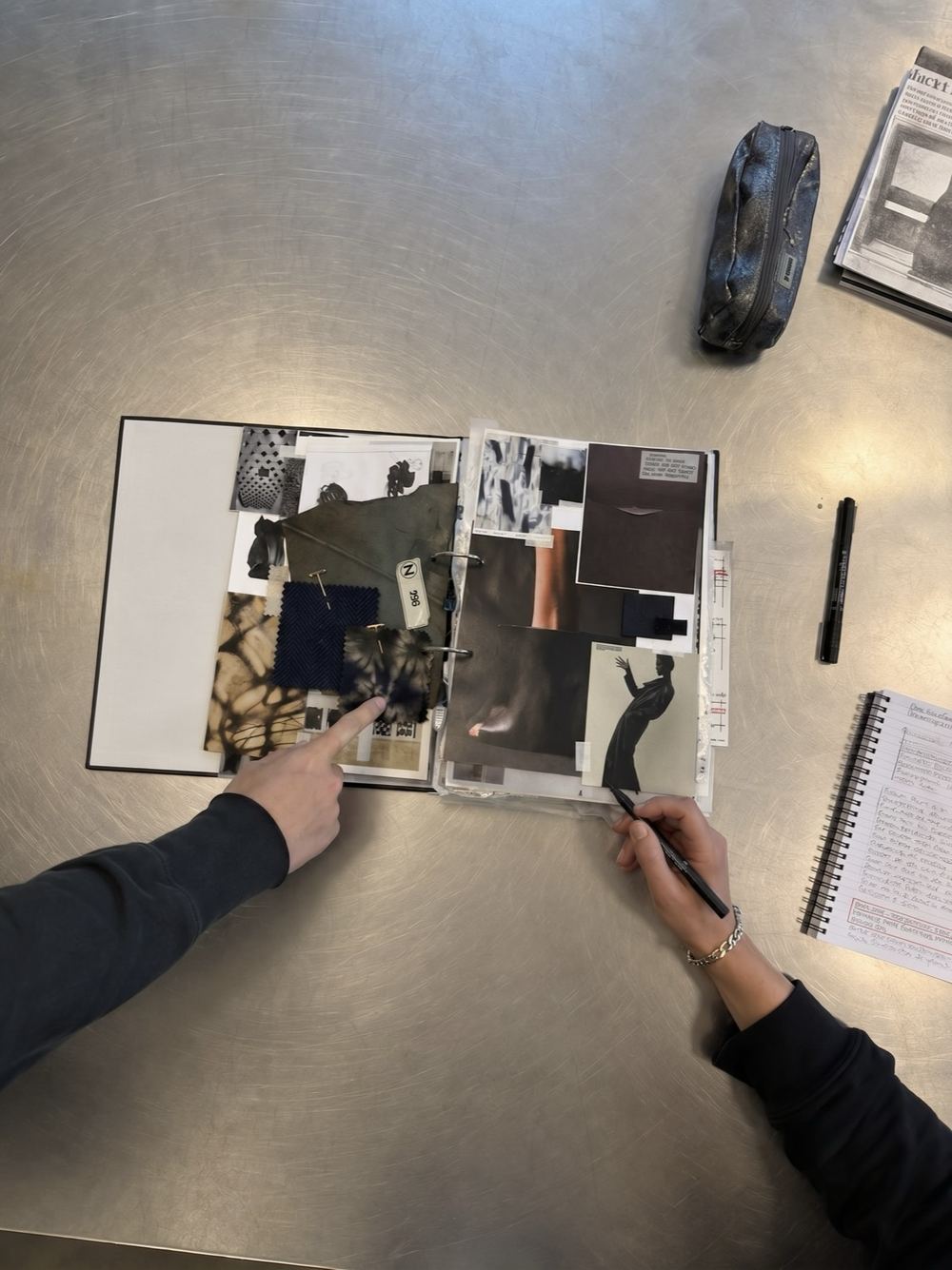 A one-to-one portfolio critique: two people reviewing a sketchbook of research, fabric swatches and design references on a studio table